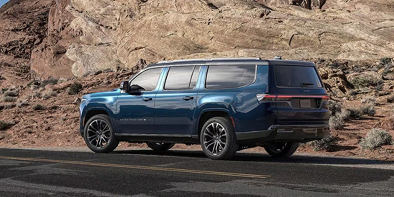 The image features a blue Jeep Wagoneer, captured in a rugged desert landscape, emphasizing its premium design and off-road capability with a striking backdrop of rocky terrain.
